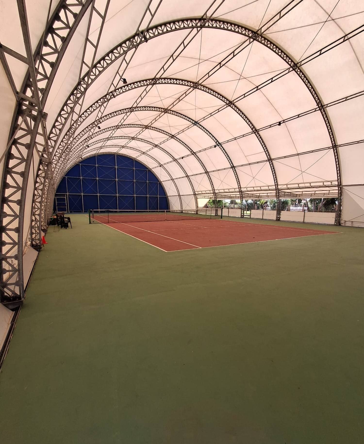 Campo da tennis rosso sotto la tensostruttura bianca, con archi in acciaio e parete di fondo blu